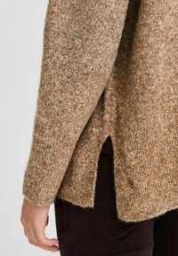 Brauner Wollpullover mit strukturierter Oberfläche, seitlichem Schlitz und Rippbündchen, kombiniert mit dunkelbraunen Hosen.
