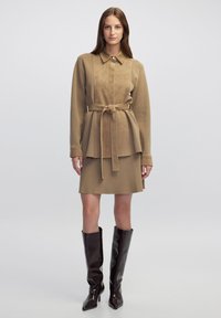 Vestido marrón claro con cinturón y cuello, que presenta mangas largas y un diseño en capas, combinado con botas de charol marrones oscuras hasta la rodilla. Textura suave.