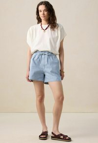 Top blanc à manches courtes, shorts rayés bleus avec une taille élastique, et sandales marron à brides croisées. Tissu lisse avec une coupe décontractée.