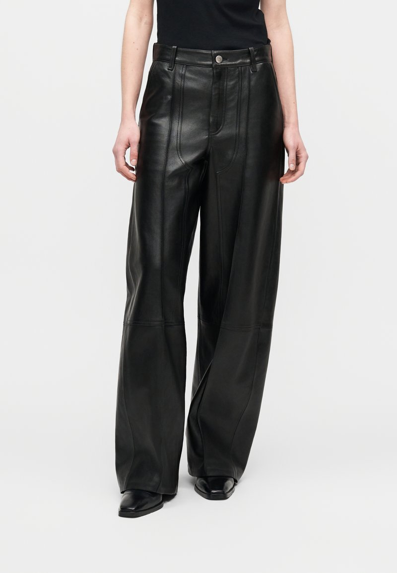 Γυναίκα με ψηλόμεσα, φαρδιά μαύρα δερμάτινα παντελόνια με ραμμένες λεπτομέρειες πάνελ και μαύρα μυτερά παπούτσια.