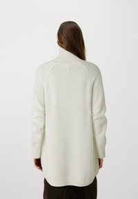 Un long pull en ivoire en tricot doux avec un col haut, des poignets côtelés et un ourlet incurvé, présentant un design de raglan sans couture.