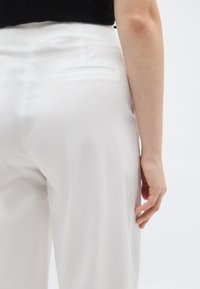 Άτομο που φοράει λευκό, ραμμένο παντελόνι και μαύρο τοπ, στέκεται με το δεξί χέρι χαλαρό στο πλάι, μπροστά σε απλό φόντο.