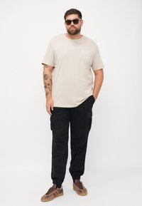 Beige katoenen t-shirt met een klein logo, gecombineerd met zwarte cargobroek met zijzakken, en bruine sneakers met een gestructureerde bovenkant.