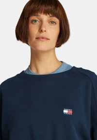 Felpa blu navy con colletto tondo e polsini a costine. Presenta una piccola patch del logo Tommy Jeans in rosso, bianco e blu. Texture morbida.