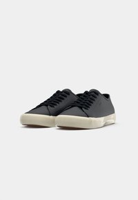 Paire de baskets basses noires avec des lacets noirs et des semelles en caoutchouc blanches sur un fond gris clair uni.