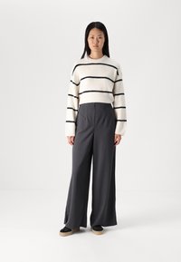 Pull en laine crème duveteux avec des rayures noires, associé à un pantalon large gris foncé. Le modèle porte des baskets noires avec une semelle beige.