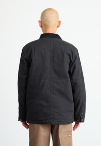 Bald person wearing a black jacket and brown pants stands facing away against a plain white background.