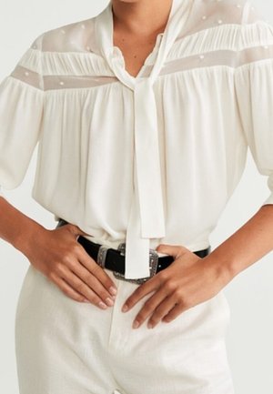 Mujer con blusa blanca con canesú transparente, mangas abullonadas y lazo en el cuello, metida en pantalones blancos con cinturón negro y hebilla plateada.