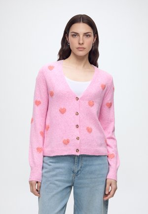 Frau trägt eine rosa Strickjacke mit orangefarbenen Herzmustern über einem weißen Oberteil und hellblauen Jeans, stehend vor einem schlichten Hintergrund.
