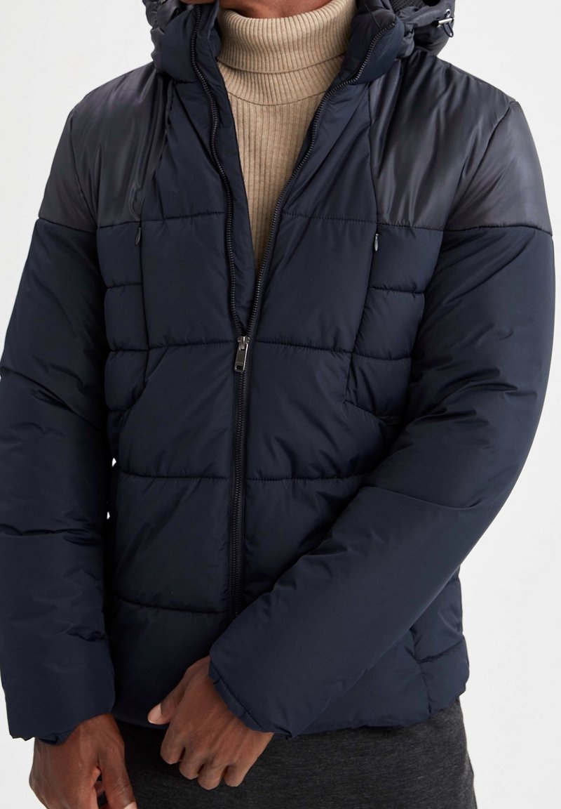 Doudoune bleu marine avec fermeture éclair devant, texture matelassée, capuche réglable et poignets élastiques. Adaptée pour les temps froids.