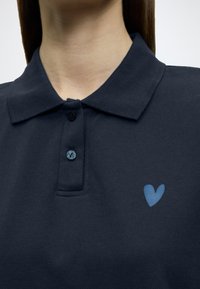 Μπλε ναυτικό πόλο με υφή υφάσματος, διαθέτει τρία κουμπιά στην εμπρός πλευρά και λογότυπο καρδιάς σε ανοιχτό γαλάζιο χρώμα στο αριστερό μέρος του στήθους.