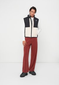 Gilet sans manches noir et crème en polaire avec fermeture éclair, associé à un pantalon large couleur rouille. Des chaussures de style sneaker noires complètent le look.