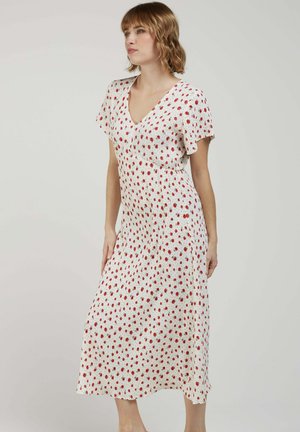 Jeune femme aux cheveux courts portant une robe midi blanche à imprimé floral rouge, manches courtes et encolure en V, se tenant devant un fond uni.