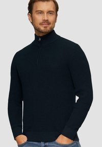 Granatowy dzianinowy sweter zapinany na zamek z wysokim kołnierzem, strukturalnym wzorem i ściągaczami na rękawach. Połączenie z jasnoniebieskimi dżinsami.