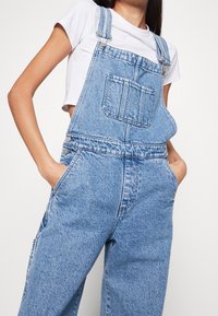Hellblaue Denim-Overalls mit weitem Bein, verstellbaren Schulterträgern und einer Fronttasche. Glatte Textur mit dezentem Verblassen.