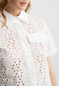 Camicia bianca a maniche corte, realizzata in tessuto ad occhiello con fori circolari, colletto con bottoni e taschino sul petto.