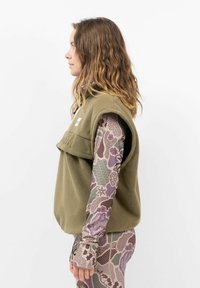 Olivgrüner Fleece-Weste mit hohem Kragen, ausgestattet mit einer Vorderklappe und Logo. Darunter ein gemusterter, langärmeliger Oberteil in Erdtönen.