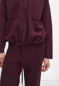 Jachetă burgundă cu fermoar, dotată cu două buzunare pe piept și un șnur în talie, asortată cu pantaloni. Țesătură moale, cu textură, și croială relaxată.