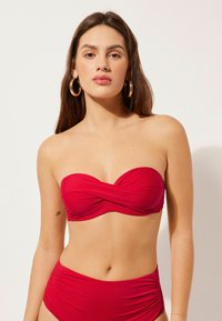 Červený bandeau bikini top s designem s otočeným předním dílem a vysokým pasem. Hladká textura tkaniny, s nařasenými detaily na bocích.