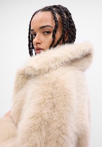Faux pelsjakke i lys beige med en myk tekstur. Har en bred, myk hette og en ren silhuett.