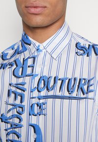 Kortärmad skjorta med knappar i vitt med blå vertikala ränder och grafisk text i svart och blått, med klassisk krage och slät textur.