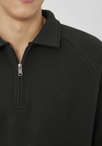 Homme portant un pullover noir à col zippé, partiellement zippé, montrant le bas du visage et le cou contre un fond uni.