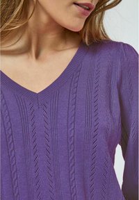 V veida purpura džemperis ar teksturētām virvju adījuma kaudzēm, ribotām malām un mīksta auduma apdari. Tuvplāns uzsver detaļas.