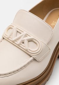 Gros plan sur un mocassin en cuir beige avec un détail de logo en relief sur la bride et une semelle épaisse marron.