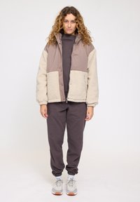 Padded jacket in beige and taupe, featuring a soft textured fabric. Worn with matching dark gray sweats and light-colored sneakers.