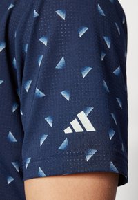 Námornícka modrá športová košeľa s textúrovanou tkaninou, ktorá obsahuje modré trojuholníkové vzory a biely logotyp Adidas na rukáve.