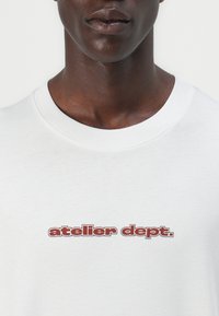 Camiseta blanca de algodón con un gráfico "atelier dept." en letras rojas, cuello redondo, mangas cortas y una textura suave.