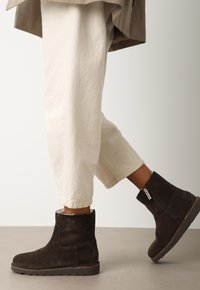 Botas de tobillo de ante marrón con cremallera lateral, tacón bajo y parte superior texturizada, combinadas con pantalones blancos enrollados.