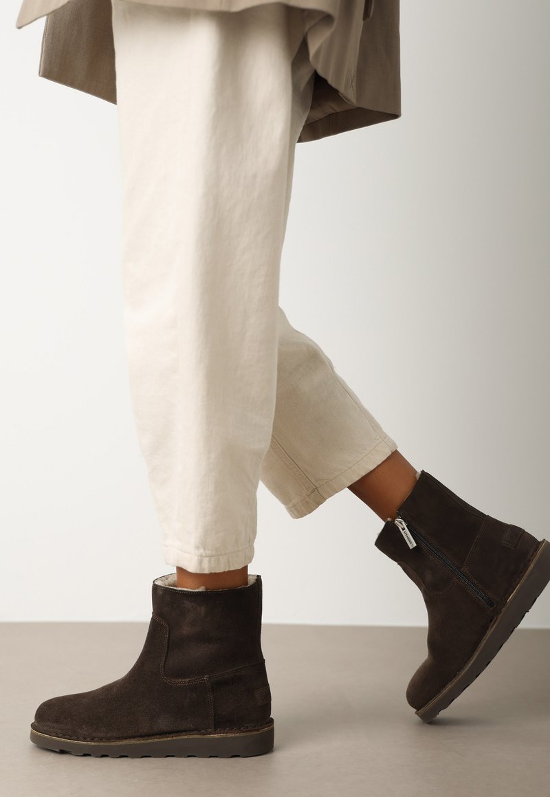 Botas de tobillo de ante marrón con cremallera lateral, tacón bajo y parte superior texturizada, combinadas con pantalones blancos enrollados.