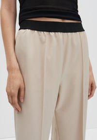 Beige wijdbenige broek met een zwarte elastische tailleband, gemaakt van een gladde, licht gestructureerde stof en een taillerende pasvorm.