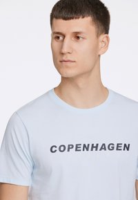 Vaaleansininen puuvillapaita lyhyillä hihoilla, jossa on rohkea musta ja kerma printti, joka sanoo "COPENHAGEN LINDBERGH" rintamalla.