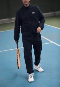 Zwart getextureerd trainingspak met ritsjack en elastische broek. Witte sneakers, terwijl er een oranje tennisracket wordt vastgehouden op een blauwe tennisbaan.
