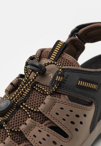 Sandalo in rete e gomma marrone con accenti neri e lacci a strisce gialle, dotato di regolatore a toggle per una vestibilità sicura. Design ventilato.