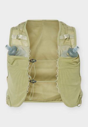 Giovane uomo con indosso una maglia nera a maniche lunghe e pantaloncini cargo oliva con un giubbotto beige per l’idratazione, che tiene due bottiglie d'acqua.