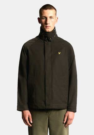 Joven con chaqueta marrón oscuro con cuello alto y logo amarillo de pájaro, combinado con pantalones verde oliva, de pie frente a un fondo liso.