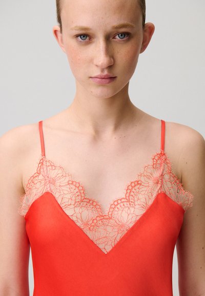 Giovane donna con occhi azzurri che indossa un top rosso con dettagli in pizzo floreale sullo scollo e spalline sottili.