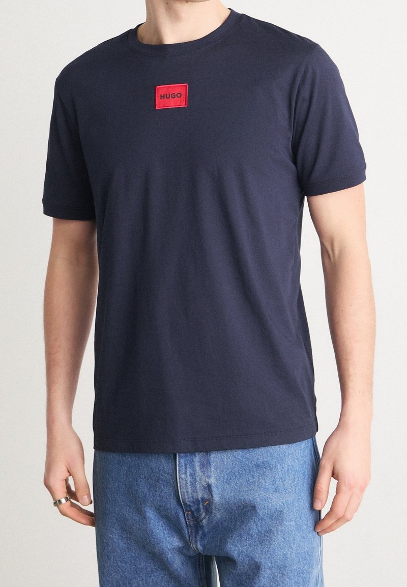 T-shirt en coton marine avec col rond et manches courtes. Présente un patch logo rouge rectangulaire "HUGO" sur la poitrine. Coupe classique.