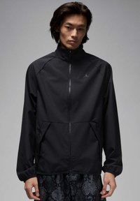 Chaqueta cortavientos negra con cuello alto, cremallera frontal y dos bolsillos laterales. Textura suave, silueta ajustada, con un detalle de logo sutil.