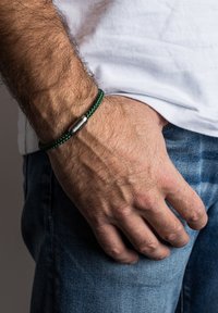 Nusivertas žalias nertas apyrankė su sidabriniu metaliniu užsegimu, dėvima ant rankos. Fone matyti balti marškiniai ir džinsinio audinio medžiaga.