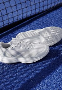 Paire de chaussures de sport blanches posées sur une surface de terrain texturée bleue près d’un filet de sport noir.