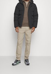 Czarna pikowana kurtka z kapturem, brązowy sweter pod spodem, beżowe spodnie oraz czarno-zielone sneakersy na białej podeszwie.