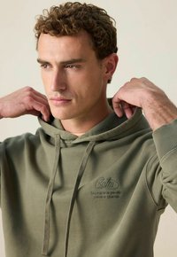 Jonge man die de capuchon van een olijfgroen sweatshirt met geborduurde tekst "Calm" op de borst aanpast, tegen een lichte effen achtergrond.