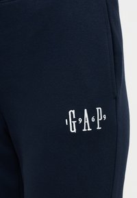 Ναυτικό μπλε ύφασμα με λευκό κεντητό κείμενο "GAP 1969" κοντά σε ραφή, υποδεικνύοντας casual ρούχα όπως φόρμες ή τζόγκερς.