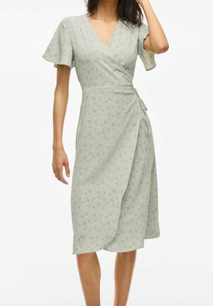 Vrouw in een lichtgroene, bloemenwrapper met korte, vloeiende mouwen, gestrikt in de taille, staand tegen een eenvoudige witte achtergrond.