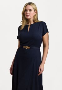 Mörkblå klänning med en nyckelhålsklack, korta ärmar och ett brunt bälte med en guldbuckla, tillverkad av ett glatt tyg med en flytande design.