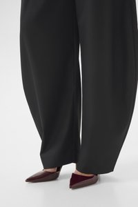 Wide black trousers paired with pointed-toe, glossy dark burgundy high heels on a white floor.